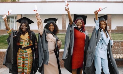 Group of young afro american female student dressed in black graduation gown. Campus as a background. Girls cheerfully smiling with arms up, holding diploma.